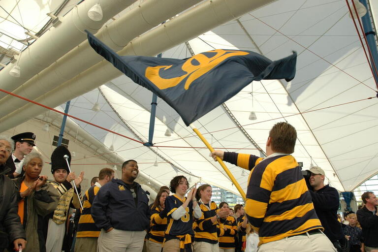 A person in a Cal rugby shirt swings a large flag with the Cal Athletics logo, people clap in the background