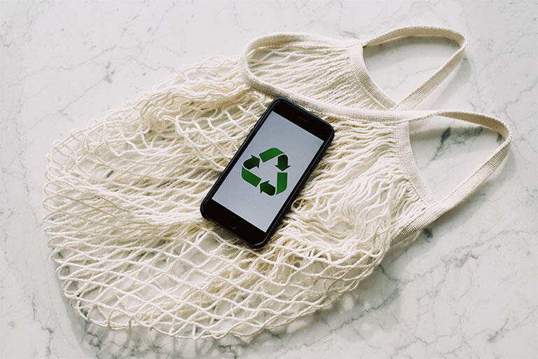 White mesh bag for shopping at the farmers market with an iPhone on top displaying the recycling symbol. 