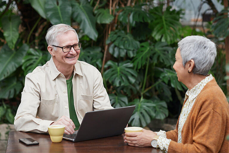 Older colleagues with grey hair sit at a table together, smiling, one on his laptop and the other holding a mug of coffee.