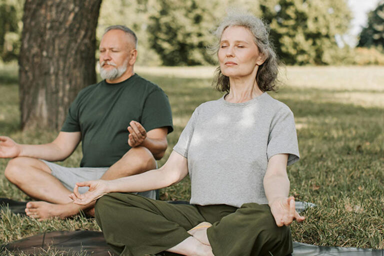 Two people sit cross-legged meditating outdoors on the grass under a tree.