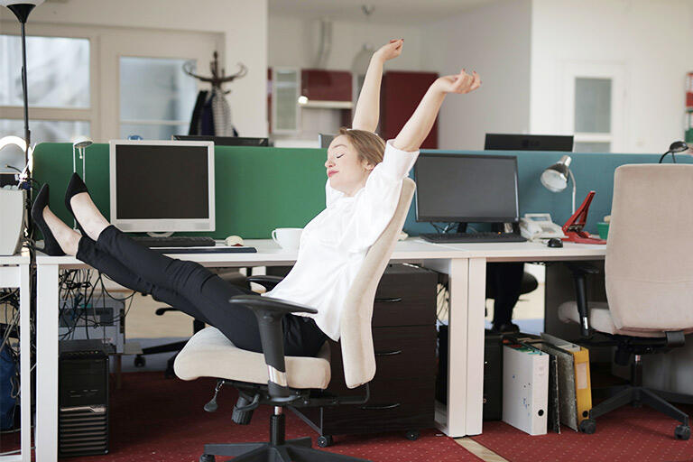 A woman sits in her chair in the office with her arms stretched up and legs out, looking very carefree