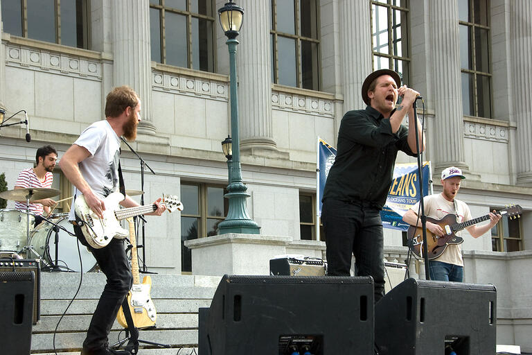 A man wearing all black sings into a microphone that he is holding with both hands, standing on the steps of Doe library. Guitarists and a drummer can be seen in the background