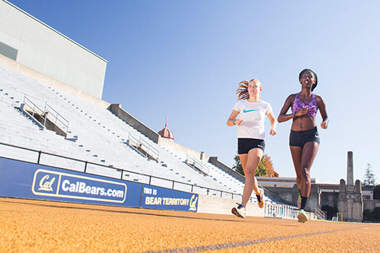 Two female students at UC Berkeley run on track with bleachers and blue sky in the background.