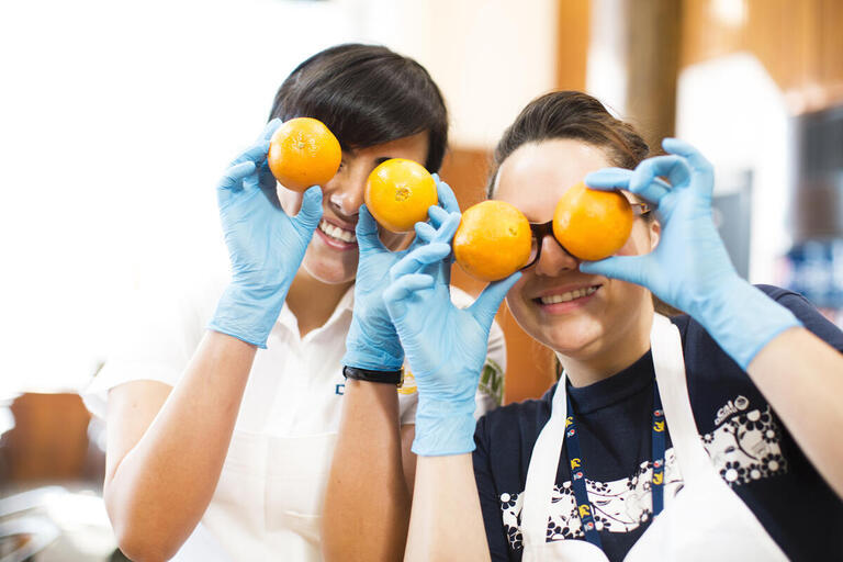 Two people wearing blue latex gloves and smiling while holding oranges in front of their eyes
