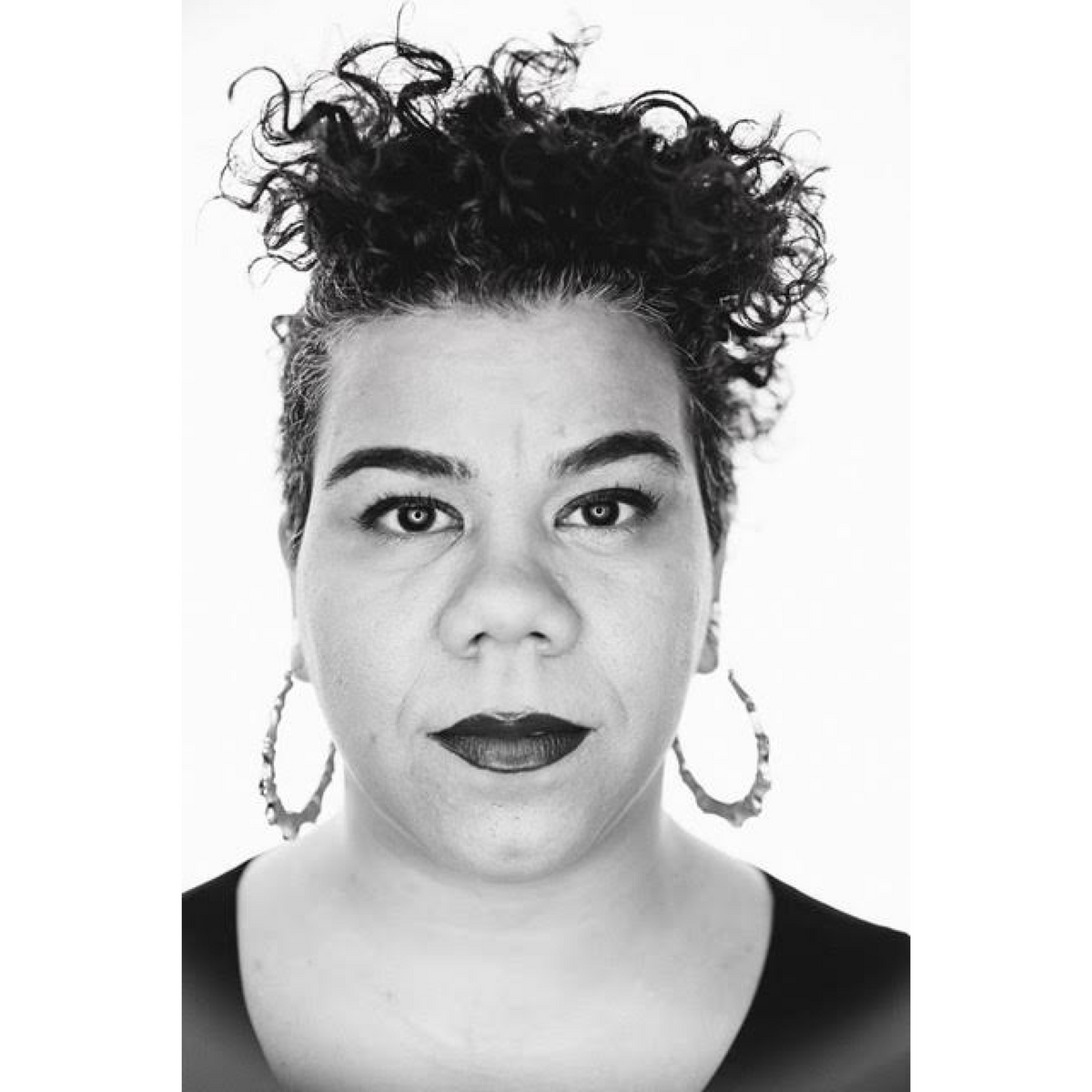 Black and white image of a woman with short dark curly hair stares into the camera. She wears large hoop earrings 