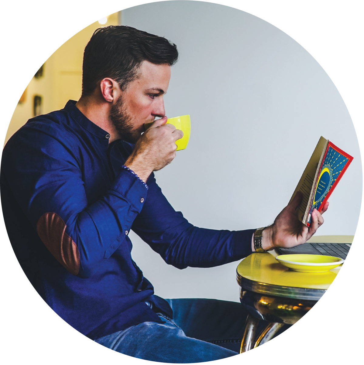 A person sitting at a table sipping from a mug and reading a book