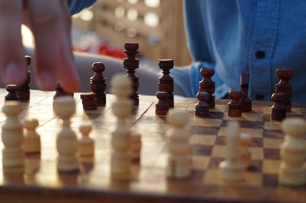 A chess board with wooden pieces in focus in the foreground, a person's shirt is visible but blurry in the background