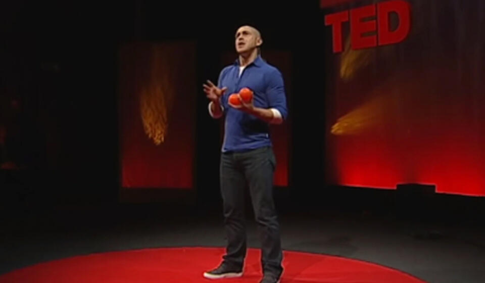 A man giving a TED talk on stage holds three juggling balls.